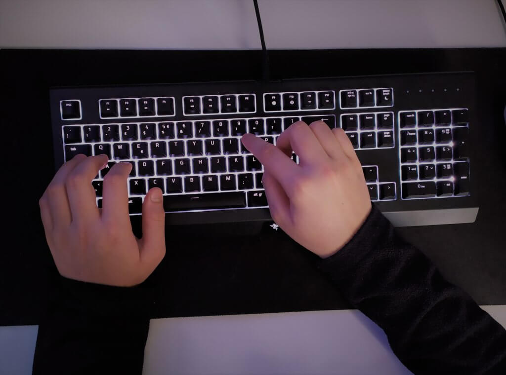 Una tastiera nera con i tasti retro illuminati da una luce bianca e la mani di un ragazzo che digitano un testo