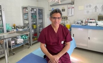 una bella immagine del dottor Alfredo Giacchetti nel suo studio medico. E’ appoggiato a un lettino da visita e indossa un camice bordò e un sorriso rassicurante.