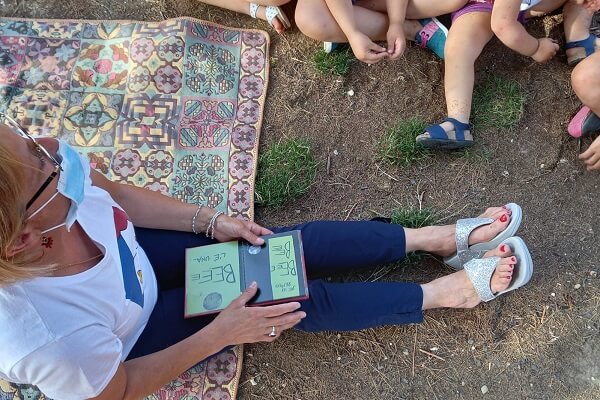Una lettrice è seduta per terra, su una coperta colorata e ha sulle gambe il libro che sta leggendo. Intorno a lei si vedono le gambine dei bimbi seduti in terra che la ascoltano