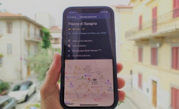 Una mano regge il cellulare collegato all'applicazione soundscape che mostra il percorso per arrivare a Piazza del Popolo, a Roma. Sullo sfondo c'è il panorama della piazza di un paese