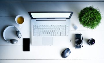 Una scrivania con un computer portatile, una tazza di caffè, una macchina fotografica, un paio di cuffie e una piantina con piccola chioma verde