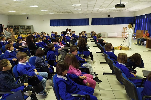 Stefania Terrè, in completo bianco, giacca e pantalone, tiene la lezione davanti a un centinaio di bambini vestiti col grembiule blu. Ai suoi piedi è accucciato Italo