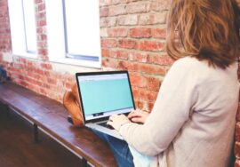 Nella foto una studentessa digita un testo al computer portatile. E' seduta su una panca addossata a una parete di mattoncini rossi. Ha le gambe allungate sulla panca e indossa jeans e una maglietta chiara. Compare di spalle