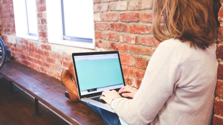 Nella foto una studentessa digita un testo al computer portatile. E' seduta su una panca addossata a una parete di mattoncini rossi. Ha le gambe allungate sulla panca e indossa jeans e una maglietta chiara. Compare di spalle