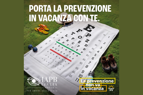 La locandina della campagna di prevenzione. Su un prato verde è adagiato un asciugamano bianco con al centro lettere e numeri che a ogni riga diventano più piccoli, come durante la visita dall'oculista. In altro la scritta: Porta la prevenzione in vacanza con te. In basso il logo di Iapb e la scritta: La prevenzione non va in vacanza. Ai bordi dell'asciugamano un paio di occhiali da sole, un paio di un paio di infradito, un cappello di paglia bianco con un nastro marrone, un paio di scarpe da trekking e una borraccia.