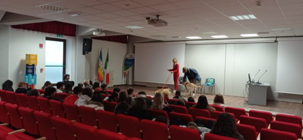 Un momento della lezione. Ripresi di spalle, gli studenti sono seduti su poltroncine imbottite rosse e seguono l'evento. In fondo alla grande sala ci sono Stefania Terrè e Mario Santoni che fanno lezione insieme ai loro cani guida