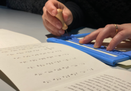 Le mani di una ragazza su una cattedra intenta a scrivere su una tavoletta Braille. Le dita sono appoggiate sulla tavoletta azzurra e la mano destra stringe un punteruolo