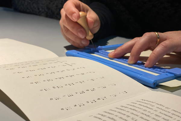 Le mani di una ragazza su una cattedra intenta a scrivere su una tavoletta Braille. Le dita sono appoggiate sulla tavoletta azzurra e la mano destra stringe un punteruolo