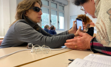 Nella foto un momento di una lezione di tecnologia assistiva. In primo piano, sedute a una scrivania, la presidente Uici di Ascoli, Gigliola Chiappini, mostra la schermata di cellulare a una socia