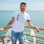 Giordano Cardellini indossa una maglietta bianca con scritte blu e jeans. È a bordo di una barca e sullo sfondo si vede il mare