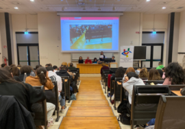 Una panoramica dell'aula universitaria in cui si è tenuto l'evento finale. Al centro la passerella e di lato le file di banchi in cui sono seduti molti studenti. In fondo, il tavolo dei relatori, a destra il grande pannello con il loro Uici Marche e in alto lo schermo gigante in cui è proiettata la sala