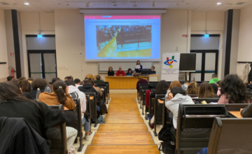 Una panoramica dell'aula universitaria in cui si è tenuto l'evento finale. Al centro la passerella e di lato le file di banchi in cui sono seduti molti studenti. In fondo, il tavolo dei relatori, a destra il grande pannello con il loro Uici Marche e in alto lo schermo gigante in cui è proiettata la sala