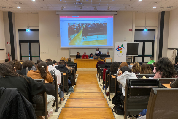 Una panoramica dell'aula universitaria in cui si è tenuto l'evento finale. Al centro la passerella e di lato le file di banchi in cui sono seduti molti studenti. In fondo, il tavolo dei relatori, a destra il grande pannello con il loro Uici Marche e in alto lo schermo gigante in cui è proiettata la sala