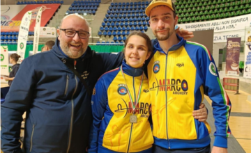 Emma Paoloni con uno splendido sorriso posa insieme al suo team al termine della bellissimo traguardo. Indossa la tuta della squadra: pantalone blu, giacchino giallo e blu con i loghi della formazione sportiva. Si trova all'interno del campo di gara