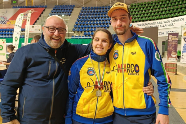 Emma Paoloni con uno splendido sorriso posa insieme al suo team al termine della bellissimo traguardo. Indossa la tuta della squadra: pantalone blu, giacchino giallo e blu con i loghi della formazione sportiva. Si trova all'interno del campo di gara